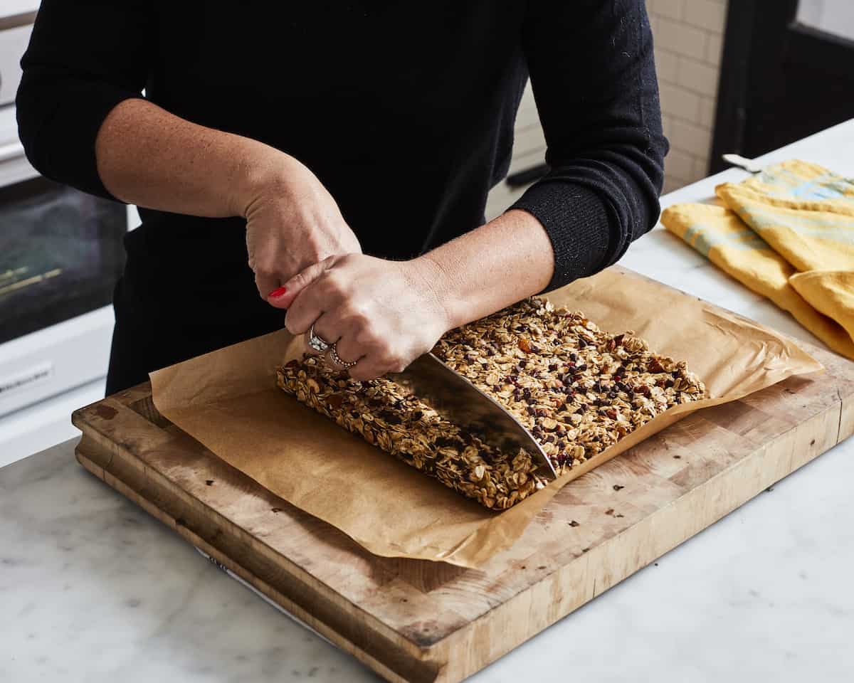 A woman with her face not visible cutting a 9x9 square of granola block placed on a parchment paper on a wooden chopping board into bars using a knife.