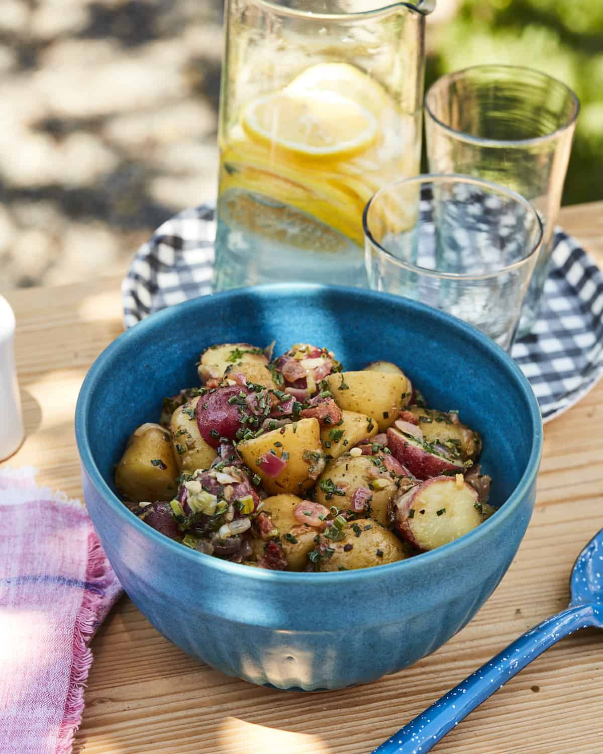 Potato salad with chives and pancetta in a blue bowl on a picnic table with gingham plates and pink linen napkins.