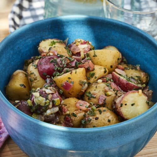 Salade de pommes de terre à la ciboulette et à la pancetta dans un bol bleu sur une table de pique-nique avec des assiettes vichy et des serviettes en lin rose