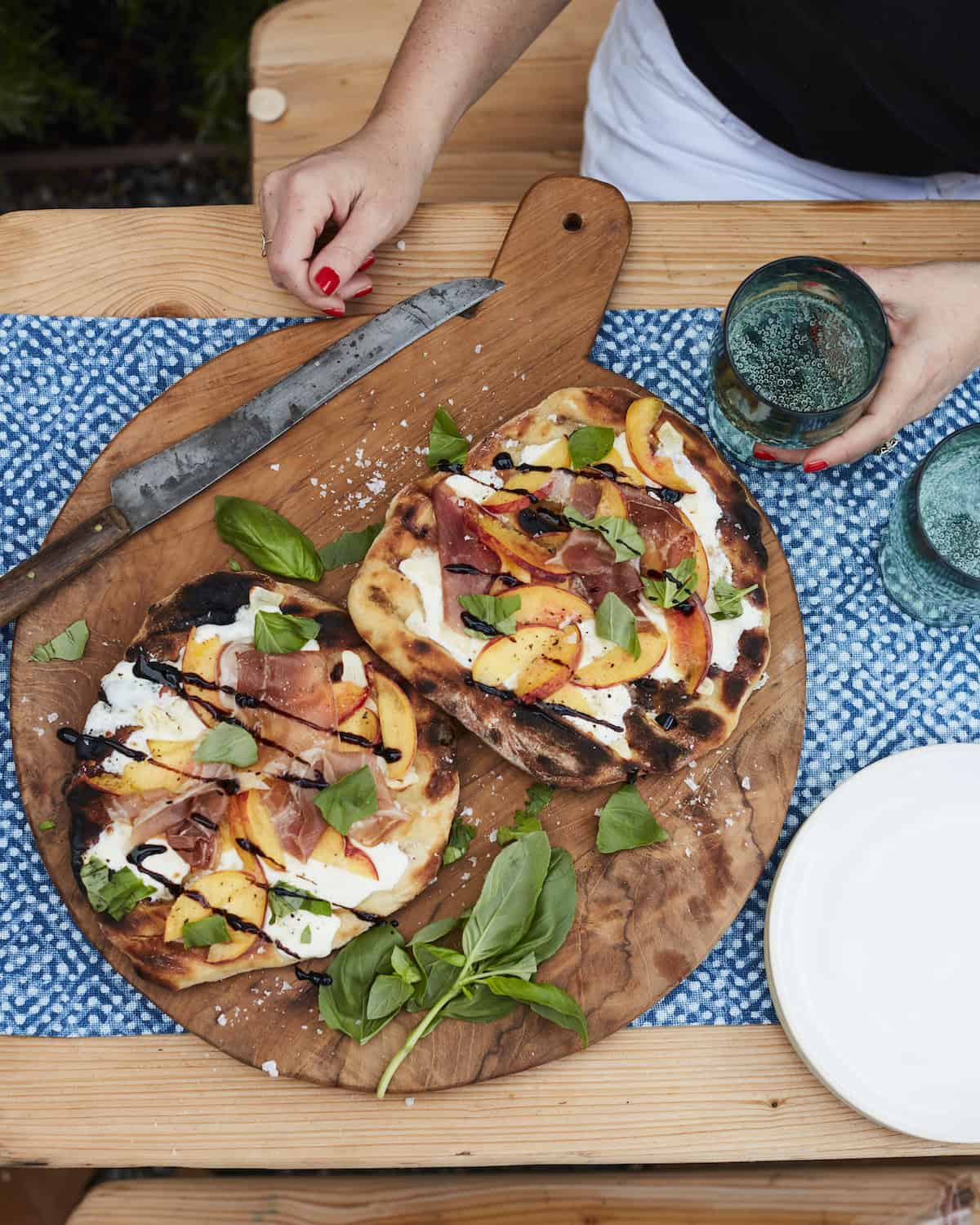 An overhead shot of a wooden board with two grilled peach and prosciutto pizzas garnished with basil along with a knife, plates and two fizzy beverages.