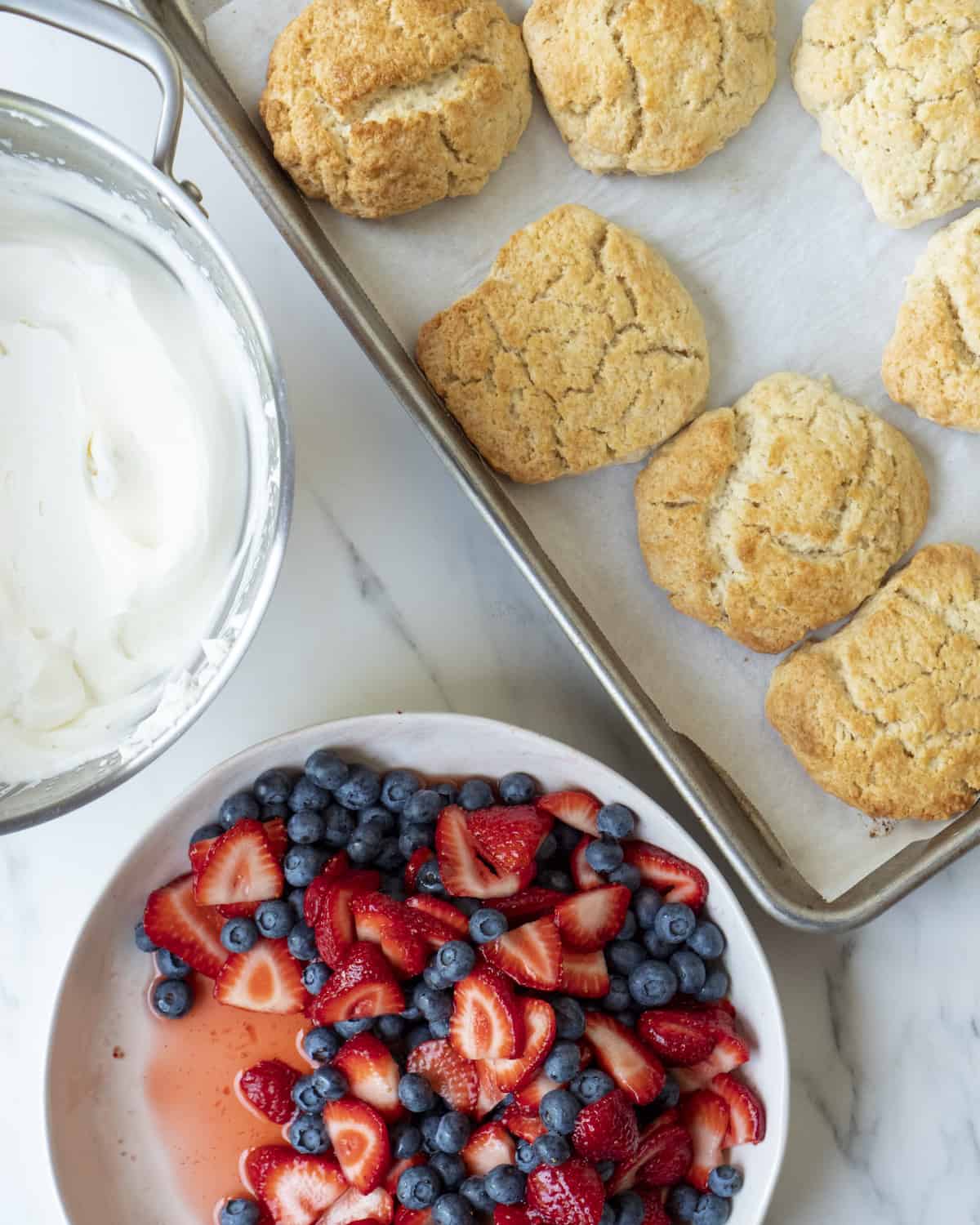 A bowl full of sugar marinated strawberries and blueberries next to a bowl full of whipped cream, next to a baking sheet with baked biscuits.  