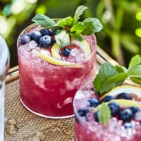 two blueberry bourbon iced lemonades in glasses garnished with mint lemon slices and blueberries on a wooden table with an ice bucket in the left corner.