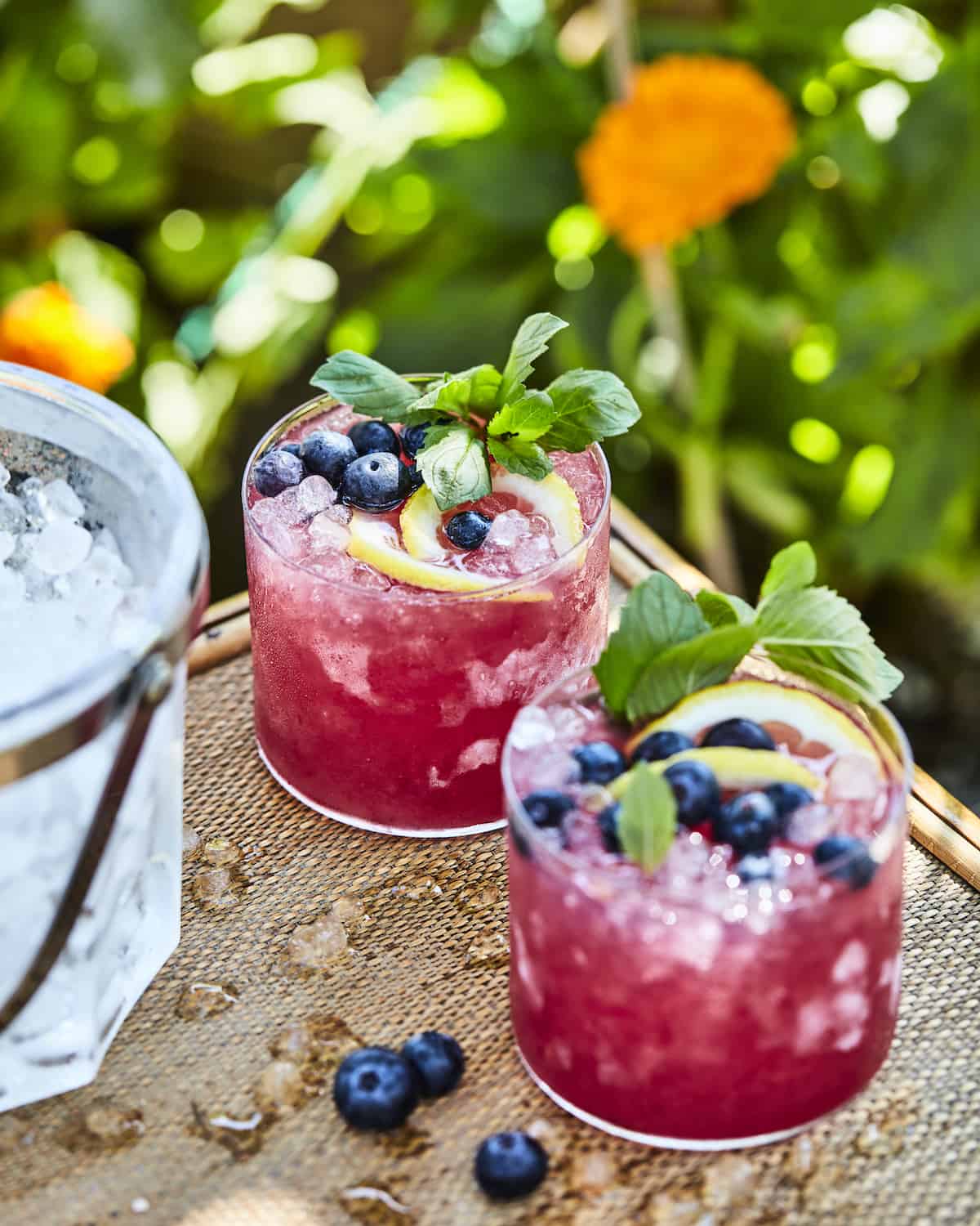 Two blueberry bourbon iced lemonades in glasses garnished with mint lemon slices and blueberries on a wooden table with an ice bucket in the left corner.
