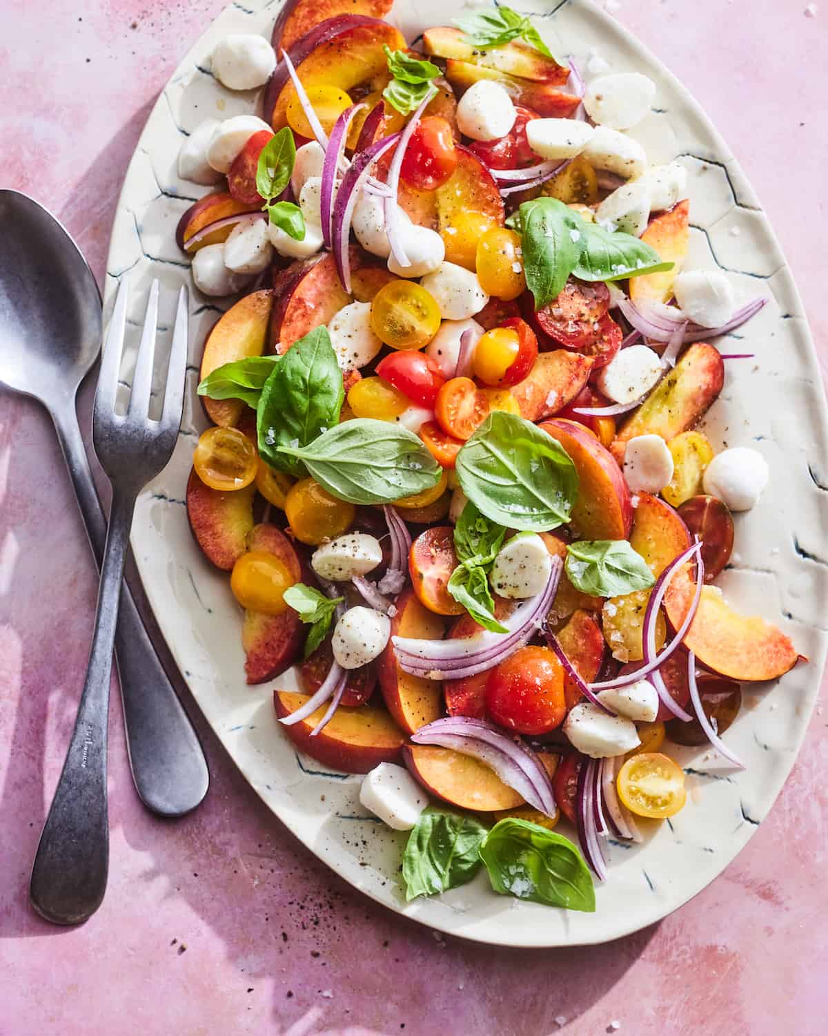 Sliced up peaches, halved cherry tomatoes, leaves of basil and sliced onions on a plate next to a serving fork and spoon.  