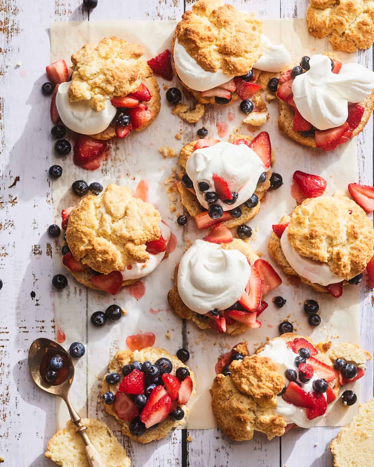 10 Strawberry Shortcakes with blueberries and whipped cream sitting on top of parchment paper on a wooden table.  