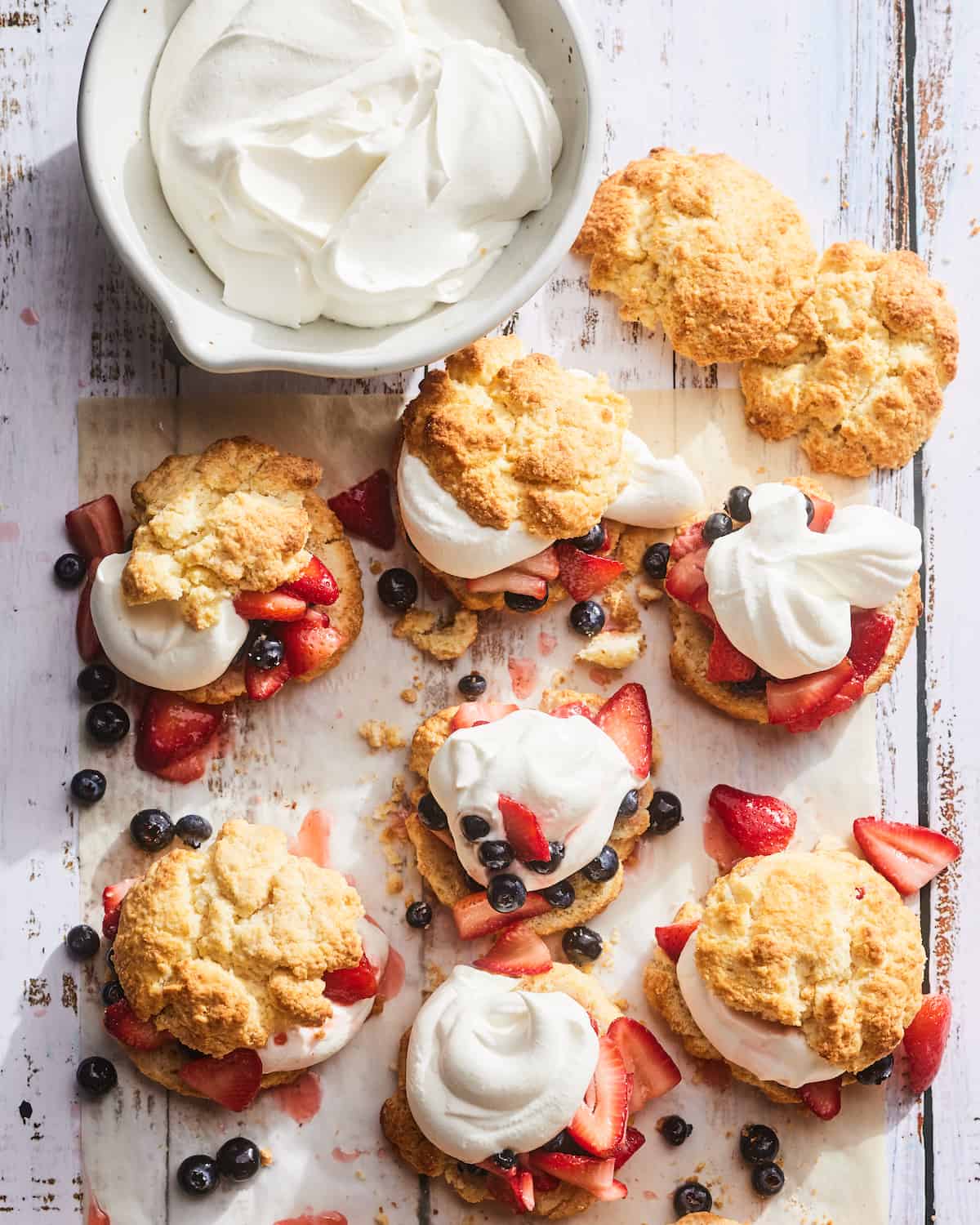 8 Strawberry shortcakes topped with whipped cream and blueberries sitting on some parchment paper next to a bowl of whipped cream.