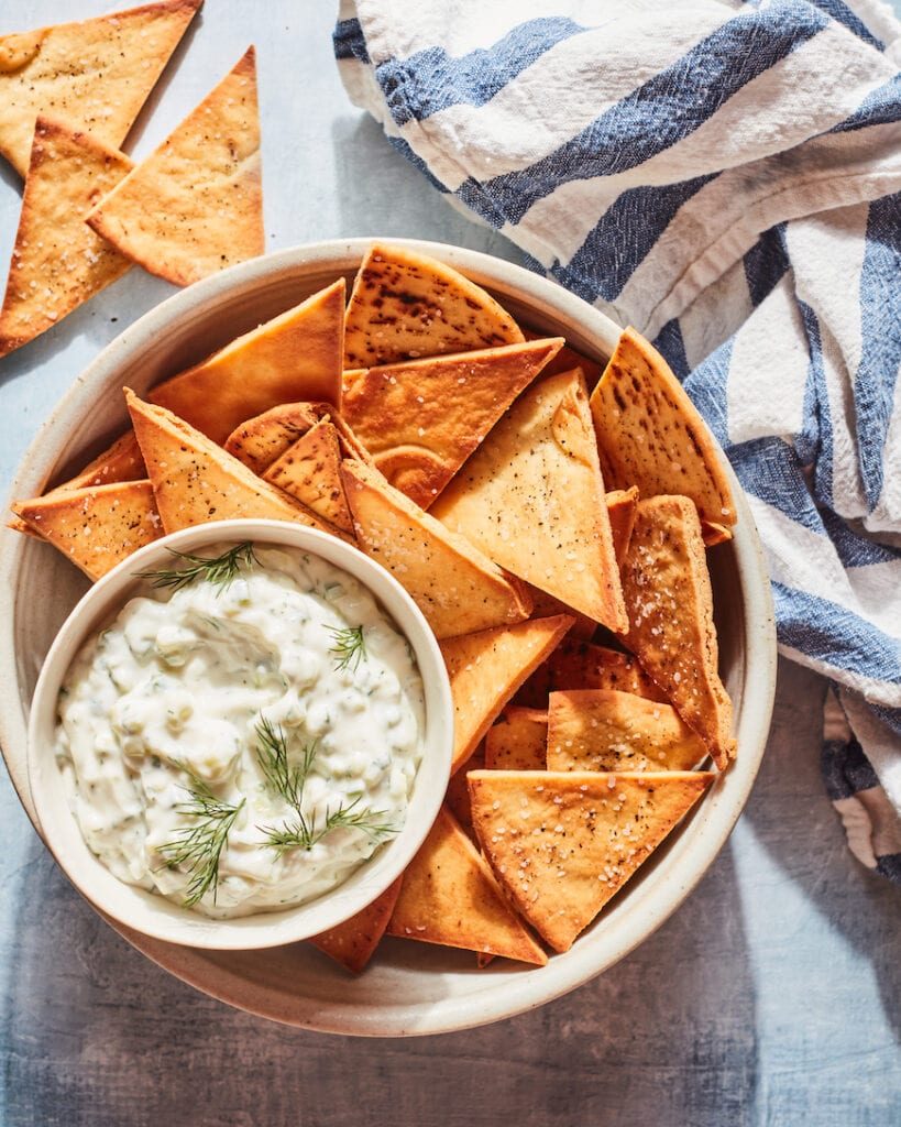 A small round bowl of homemade Tzatziki with a garnish of 4 dill sprigs nestled in the bottom left corner of a larger bowl filled with triangular pita chips on a whitewashed light blue surface with three pita chips scattered in the top left corner and a blue and white striped kitchen towel in the top right corner.