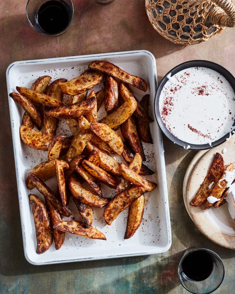 A pile of 40 -45 potato wedge fries with sumac dusted on top on a white quarter sheet pan next to a bowl of garlic feta labneh dip. 