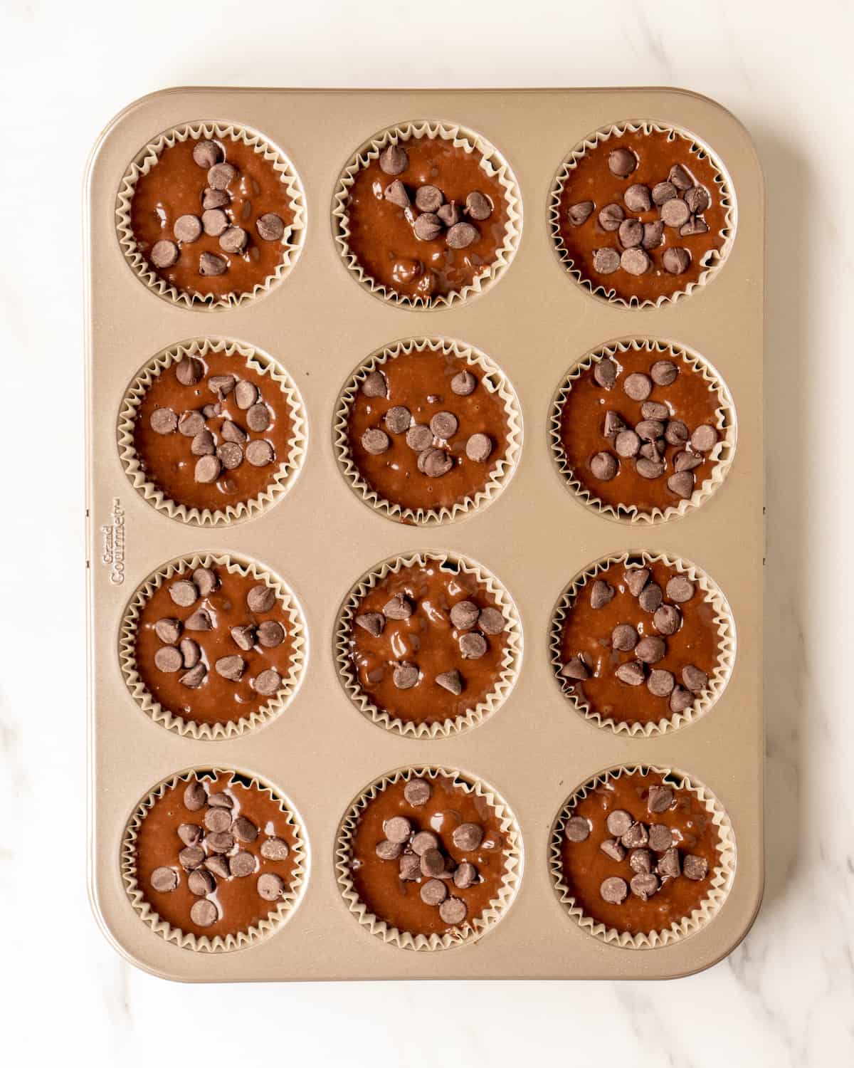 A 4x3 muffin tray lined with cupcake liners, filled with double chocolate chip muffins batter and topped with chocolate chips.