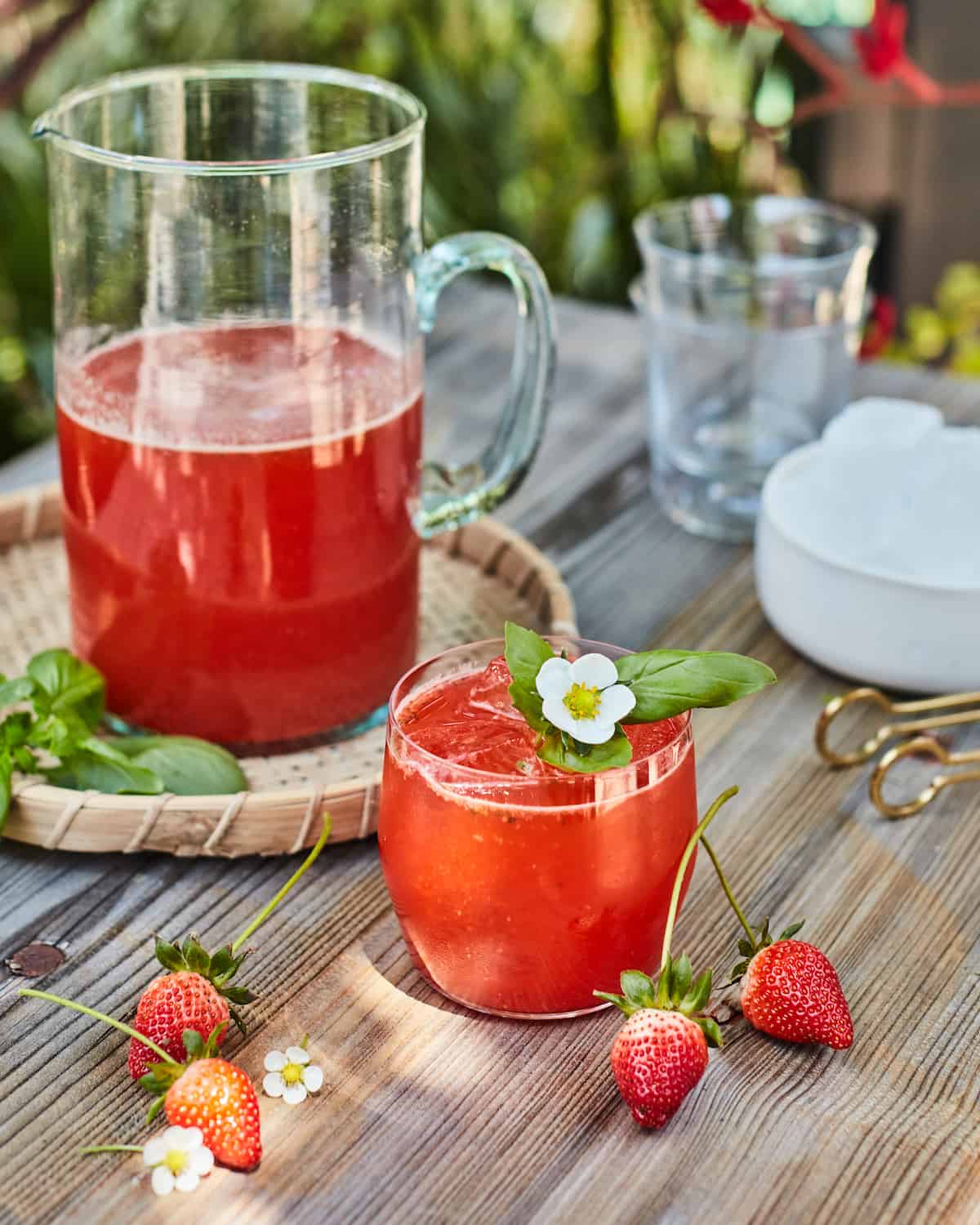 A glass of strawberry basil lemonade with a basil leaf and flower next to a pitcher of strawberry basil lemonade.