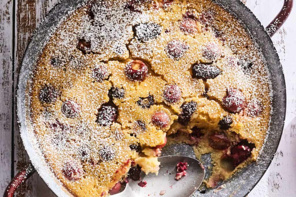 Cherry Clafoutis in a round copper metal baking dish on a white washed wood background with a dusting of powdered sugar sprinkled over top and a spoonful or two missing from the bottom right corner of the baking dish.