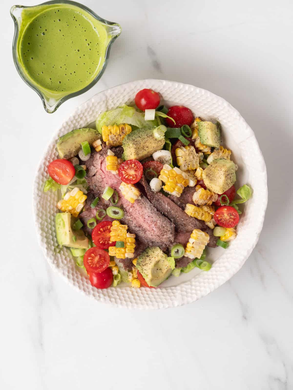 A low bowl with a grilled steak salad with avocado, corn and tomatoes, and a bowl on the side with a yogurt green goddess dressing.