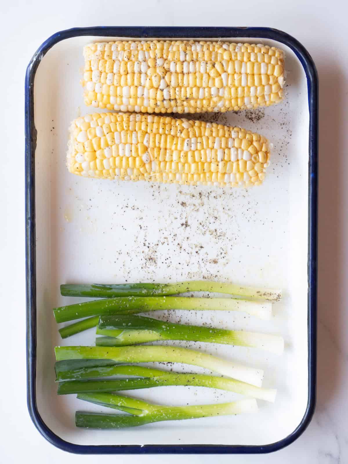 A rectangular platter with green onions and corn coated with oil and sprinkled with salt and pepper.