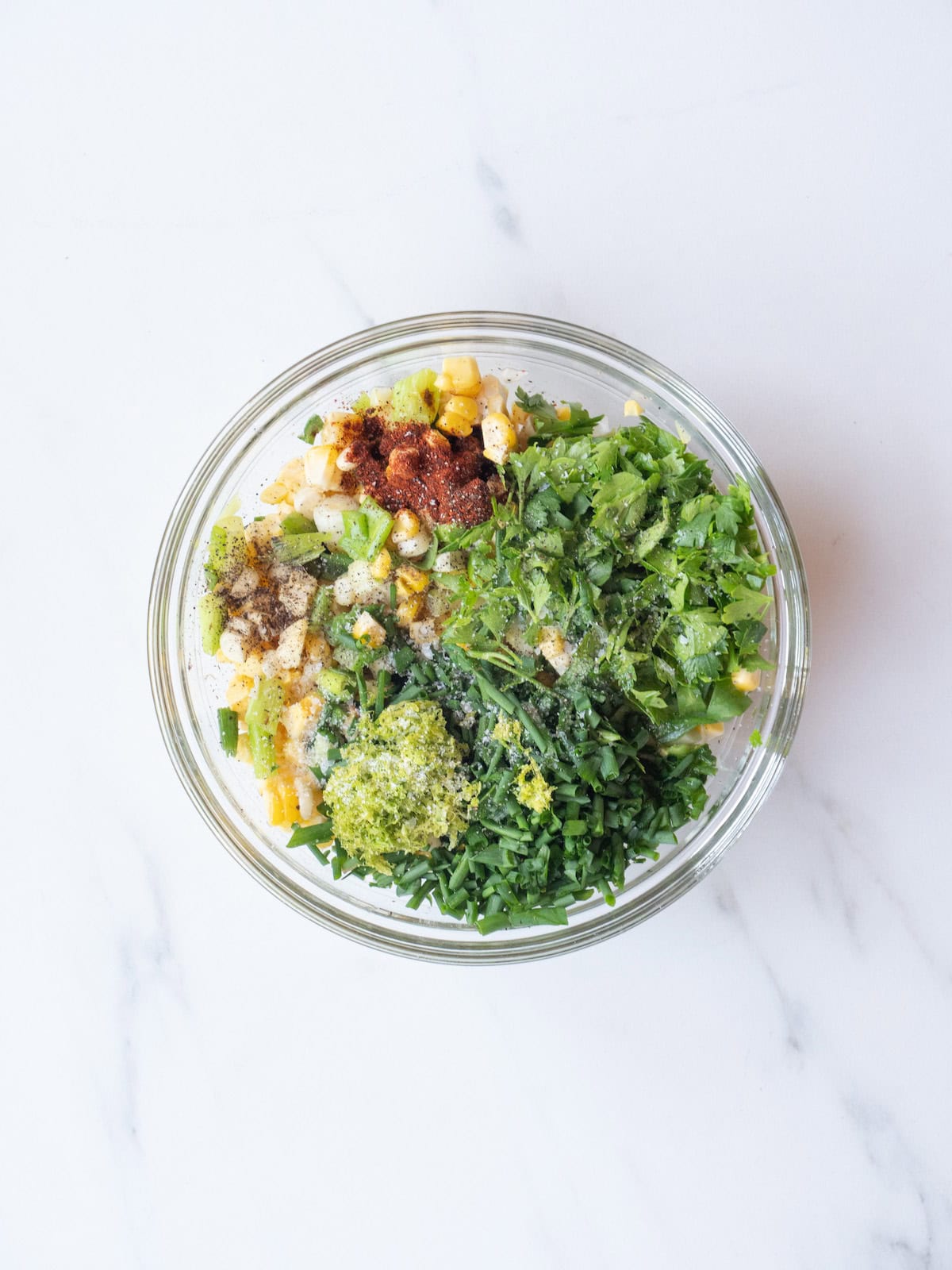 A mixing bowl with corn and green onions, with cilantro, chives, lime zest, chipotle powder, oil and lime juice.