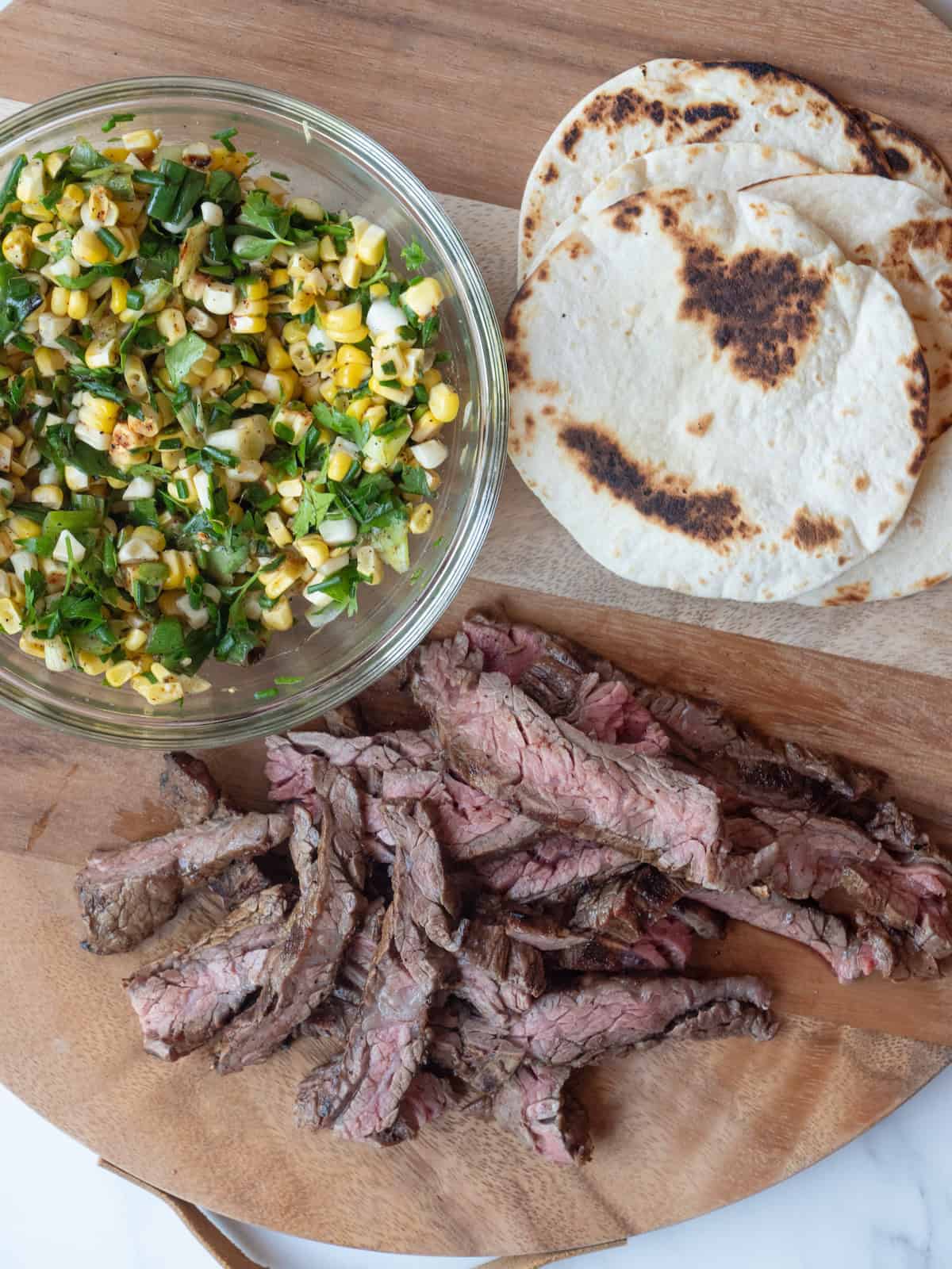 A wooden platter with charred tortillas, sliced skirt steak and a bowl with corn salsa.