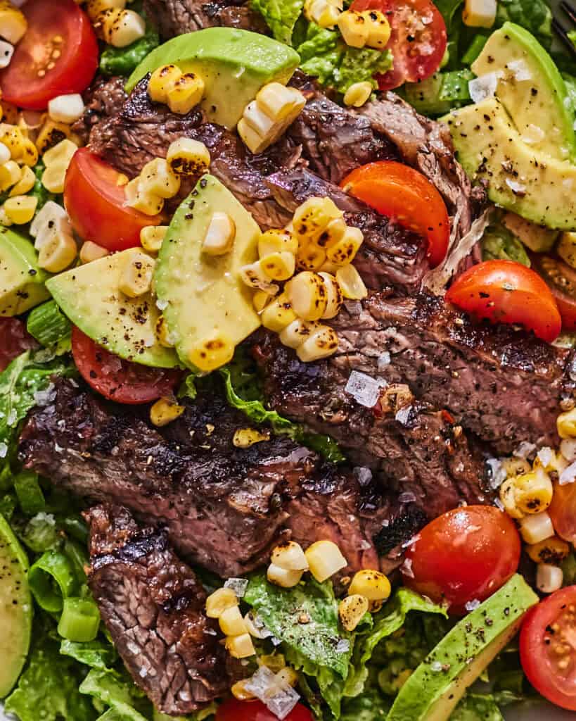 A close up shot of a grilled steak salad with tomatoes, avocado and corn.