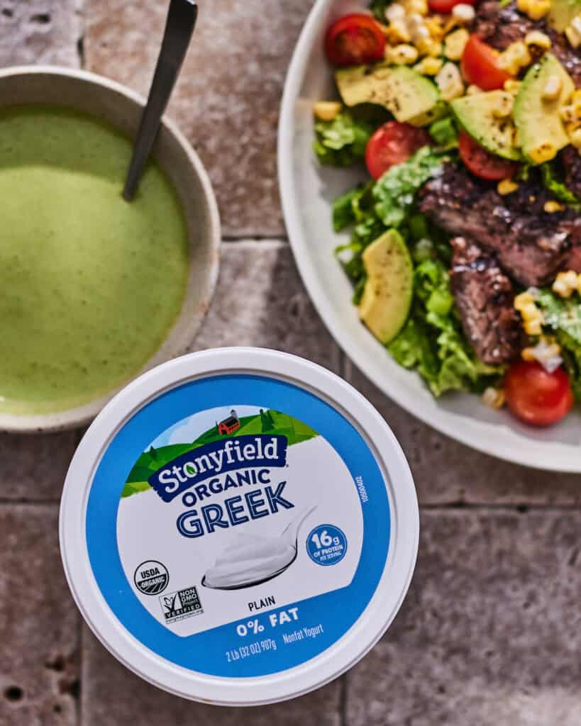 A tub of plain greek yogurt with a bowl of yogurt green goddess dressing and another bowl of a grilled steak salad.