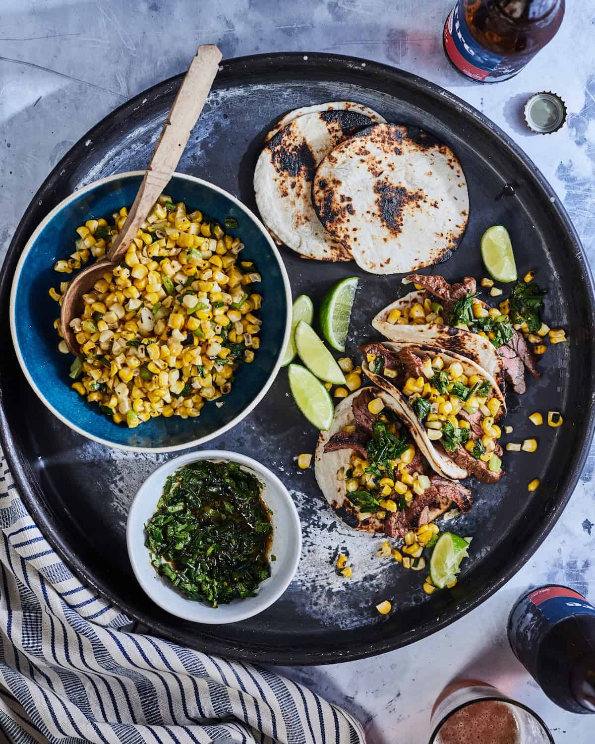 A platter with charred tortillas, a bowl with corn salsa, a bowl with cilantro relish, some wedges of lime, and some assembled tacos with steak in them.