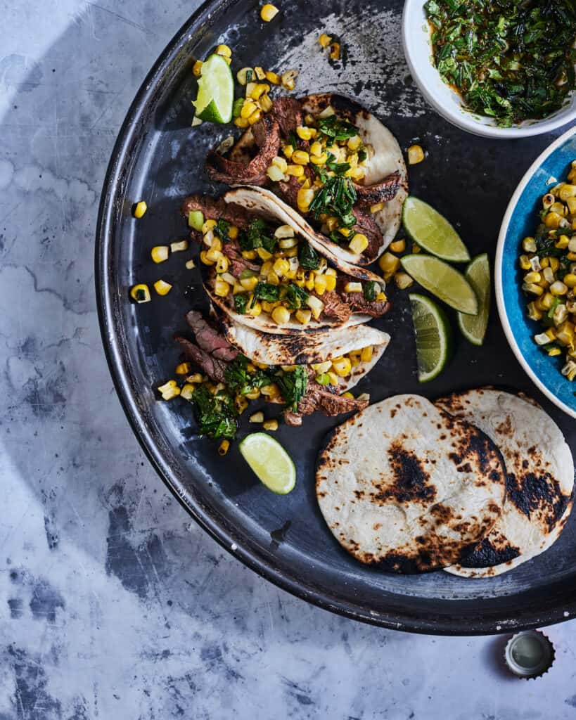 A platter with charred tortillas, a bowl with corn salsa, a bowl with cilantro relish, some wedges of lime, and some assembled tacos with steak in them.