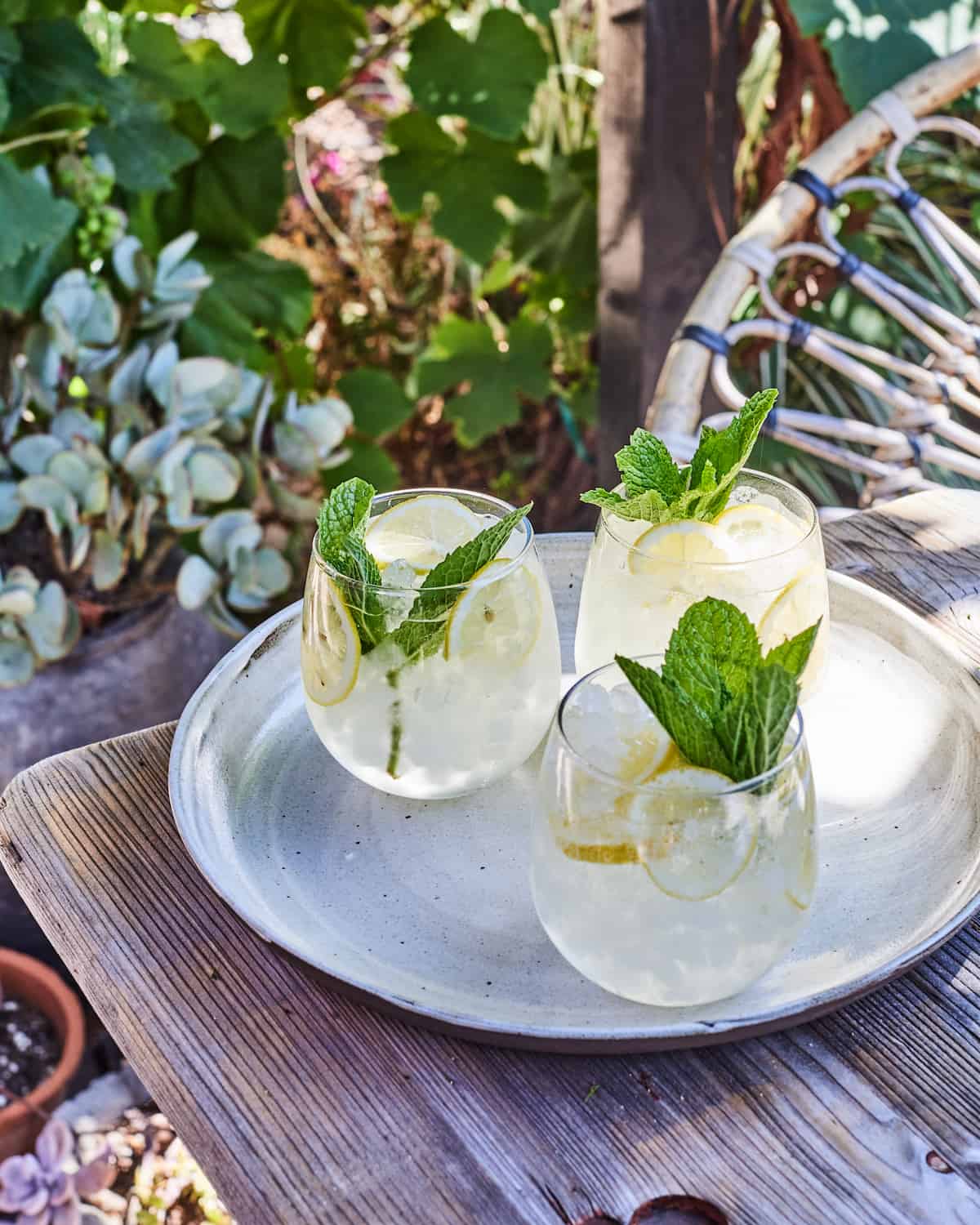 Outside on a wooden table under a lush green pergola there are three stemless wine glasses on a round metal serving tray filled with a White Wine Spritzer over ice with lemon slices and sprigs of mint for garnish. 