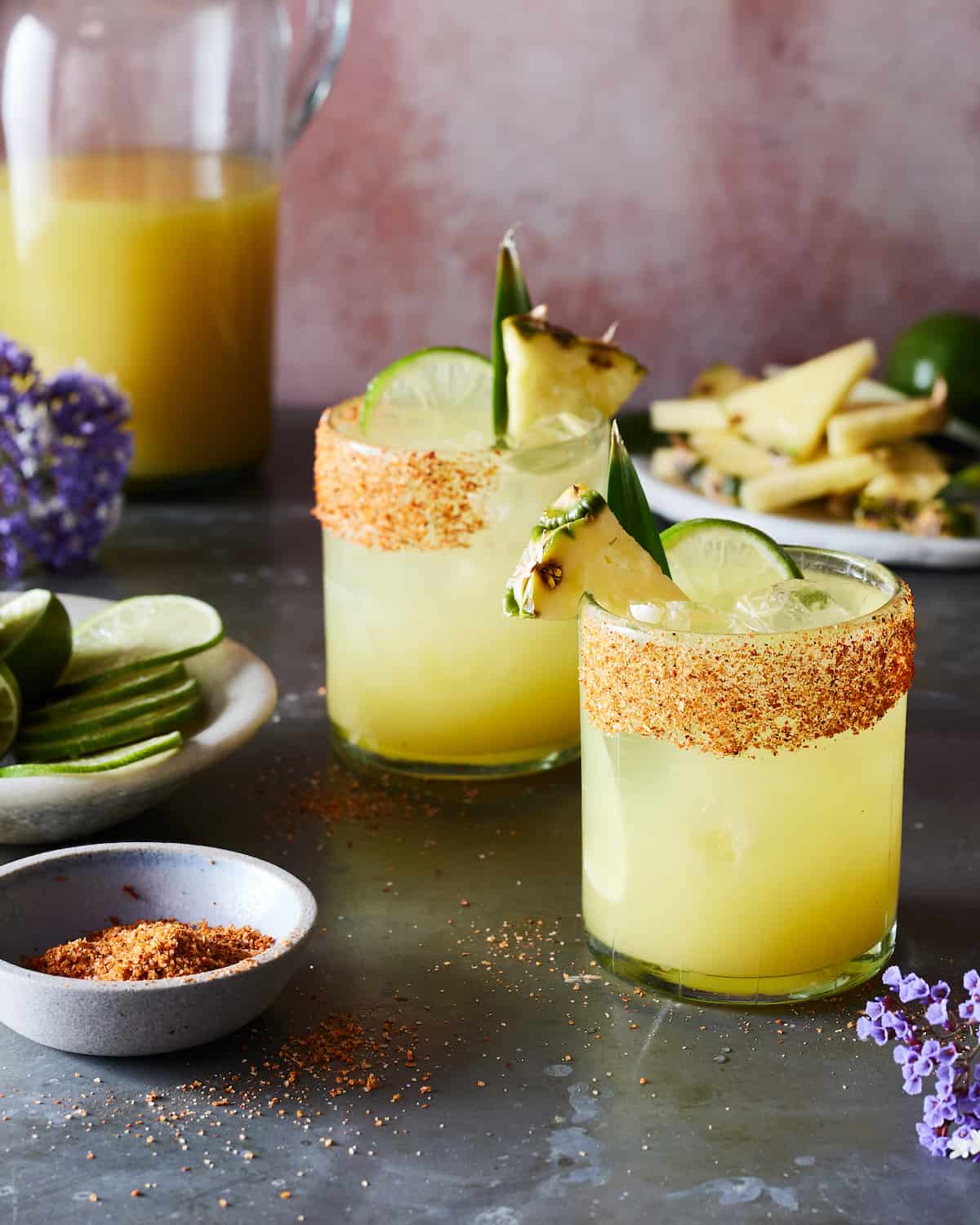 Two pineapple margaritas with Tajin lined rims with wedges of lime and pineapples attached to the rim.  