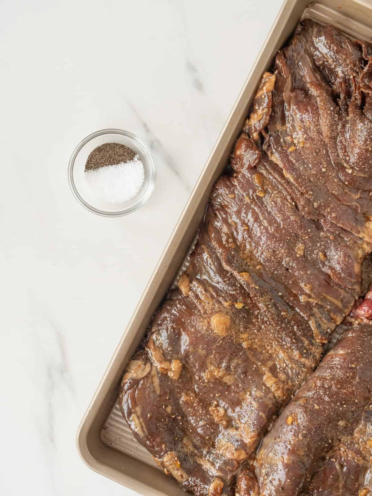 A baking sheet with skirt steak seasoned with salt and pepper, and a small mixing bowl with salt and pepper.