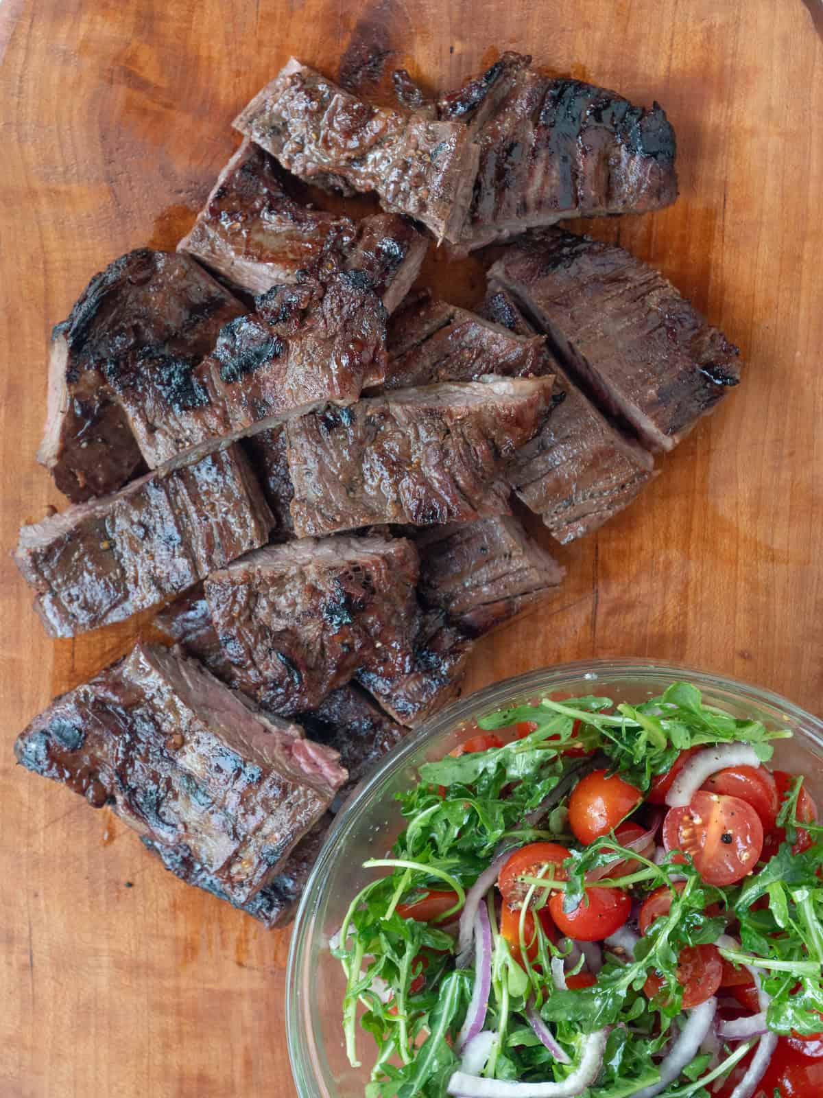 A cutting board with a chopped up skirt steak with a bowl on the side with a cherry tomato salad.