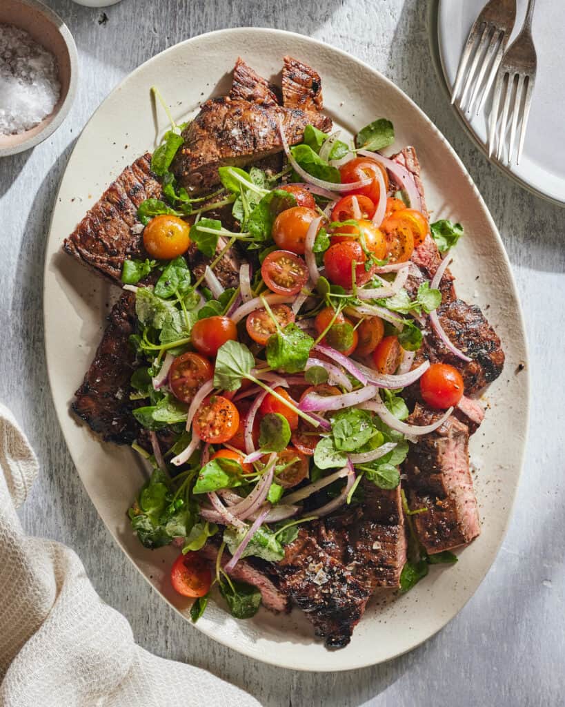 A platter with grilled skirt steak topped with a cherry tomato salad.