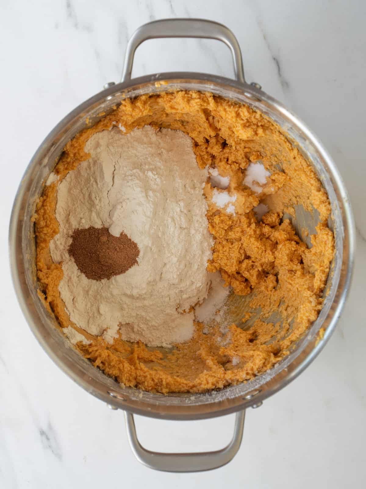 A stand mixer bowl with wet ingredients for cookies mixed together, and flour, baking soda, pumpkin spice and salt added.