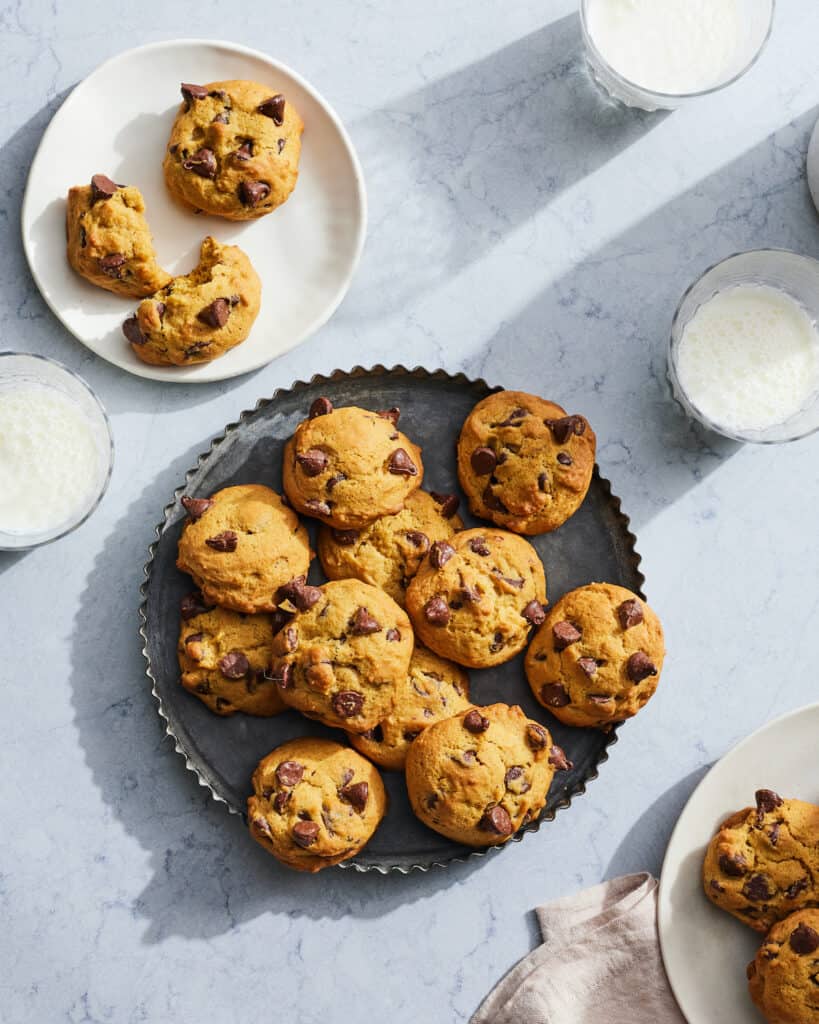 A platter with chocolate chip pumpkin cookies, along with small plates with cookies served in them, and glasses of milk.