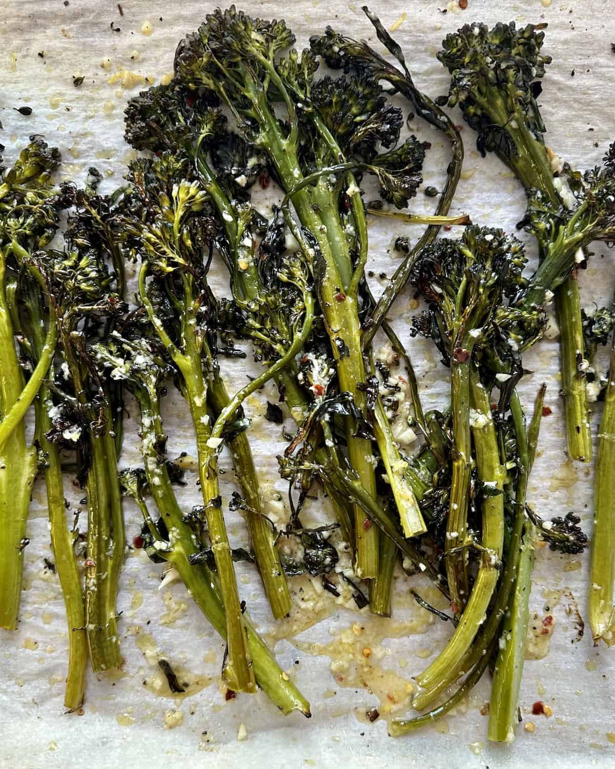A baking sheet lined with parchment paper with roasted broccolini spread evenly that has been tossed in a dressing with olive oil, minced garlic, red pepper flakes, lemon zest and juice and grated parmesan.