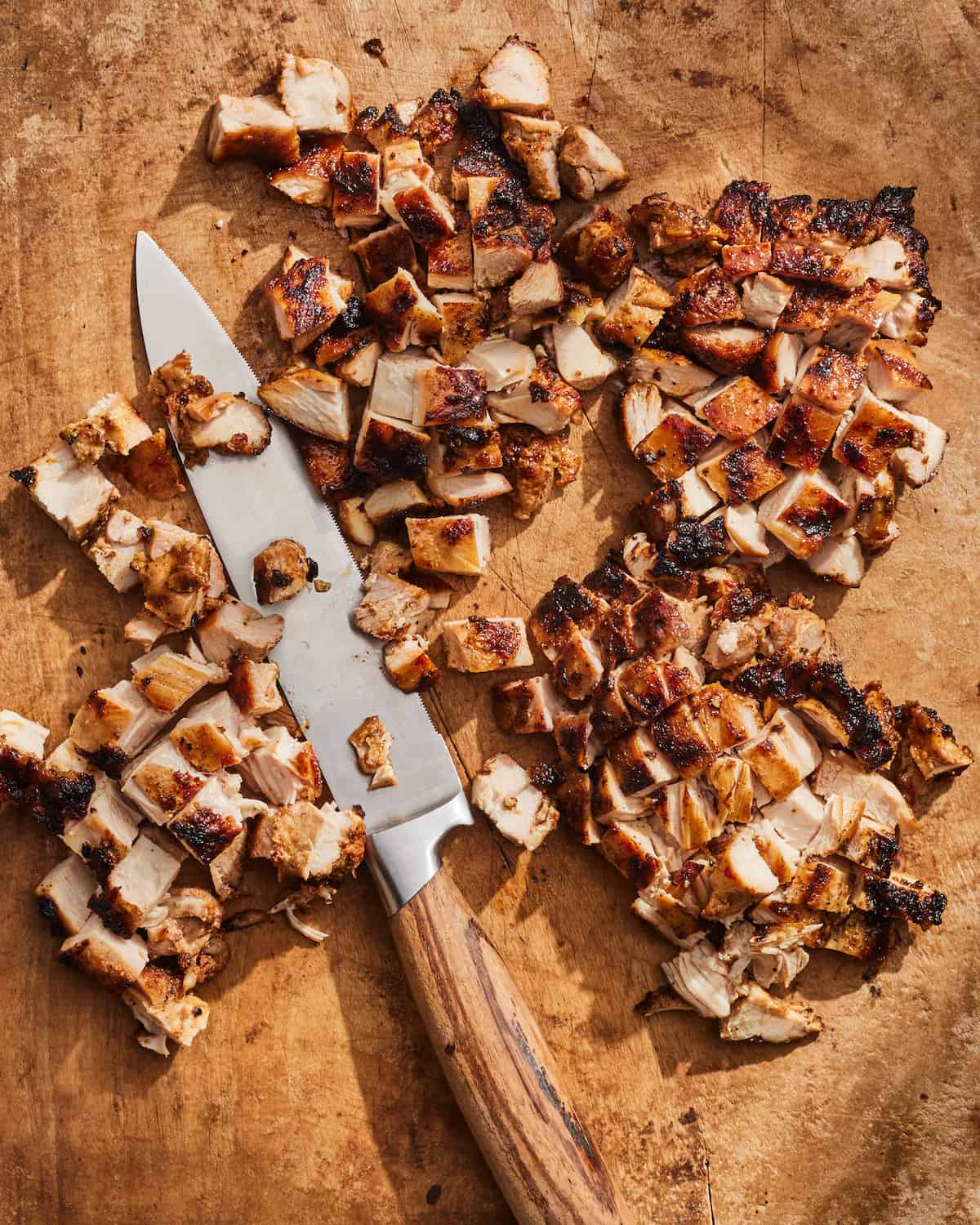 Roasted chicken that has been diced up on a wood cutting board.  A chefs knife is also laying amongst the cut up chicken on the cutting board.  