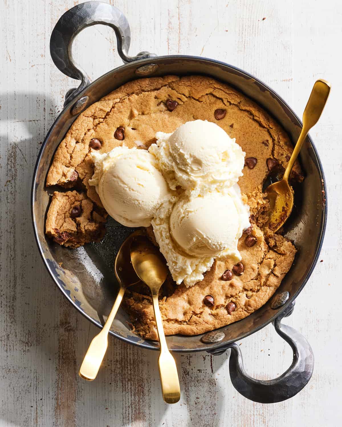 A skillet with a giant chocolate chip cookie the size of the skillet, called pizookie, topped with three scoops of vanilla ice cream, along with three spoons and few bites taken out.