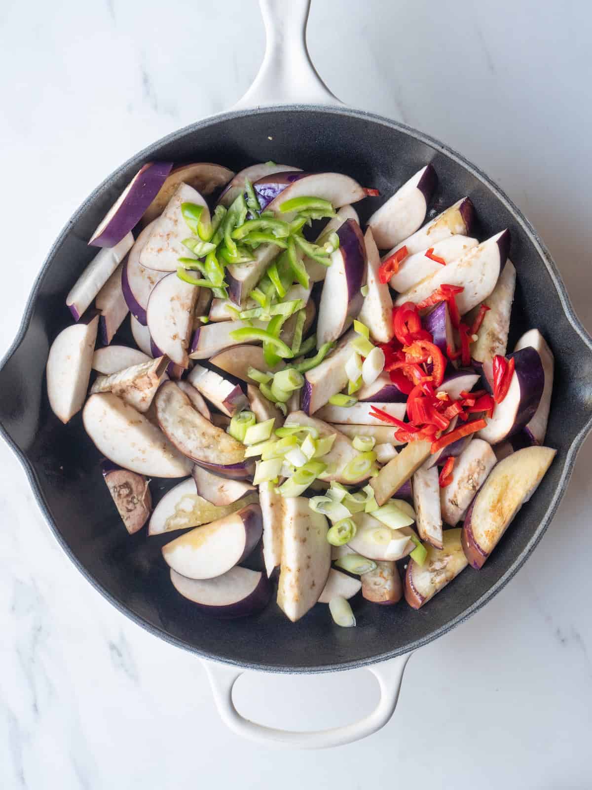 A skillet with eggplant being cooked, and peppers and green onion just added.