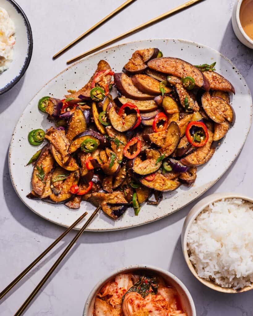 An oaval platter with spicy stir fried eggplant, along with chopsticks, a bowl of rice and a bowl of kimchi on the side.