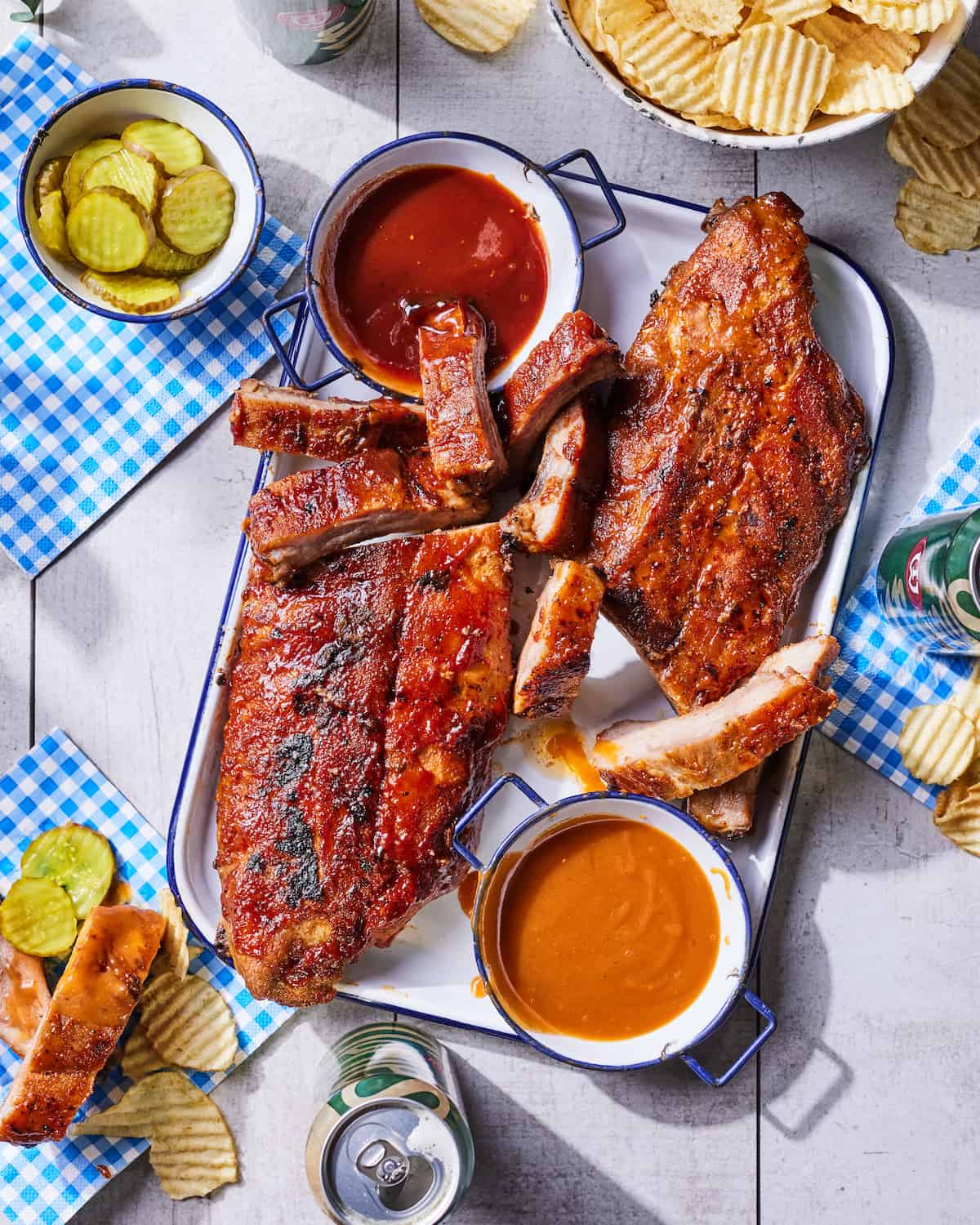 A big platter of BBQ Ribs, BBQ sauces in shallow bowls for dipping, sides of sliced pickles, and ruffle chips spread out on a white wooden table with blue and white gingham napkins