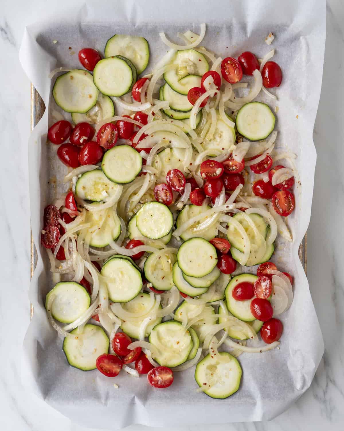 A sheet pan lined with parchment paper of sliced zucchini, yellow onion, cherry tomatoes, and drizzled with olive oil. 