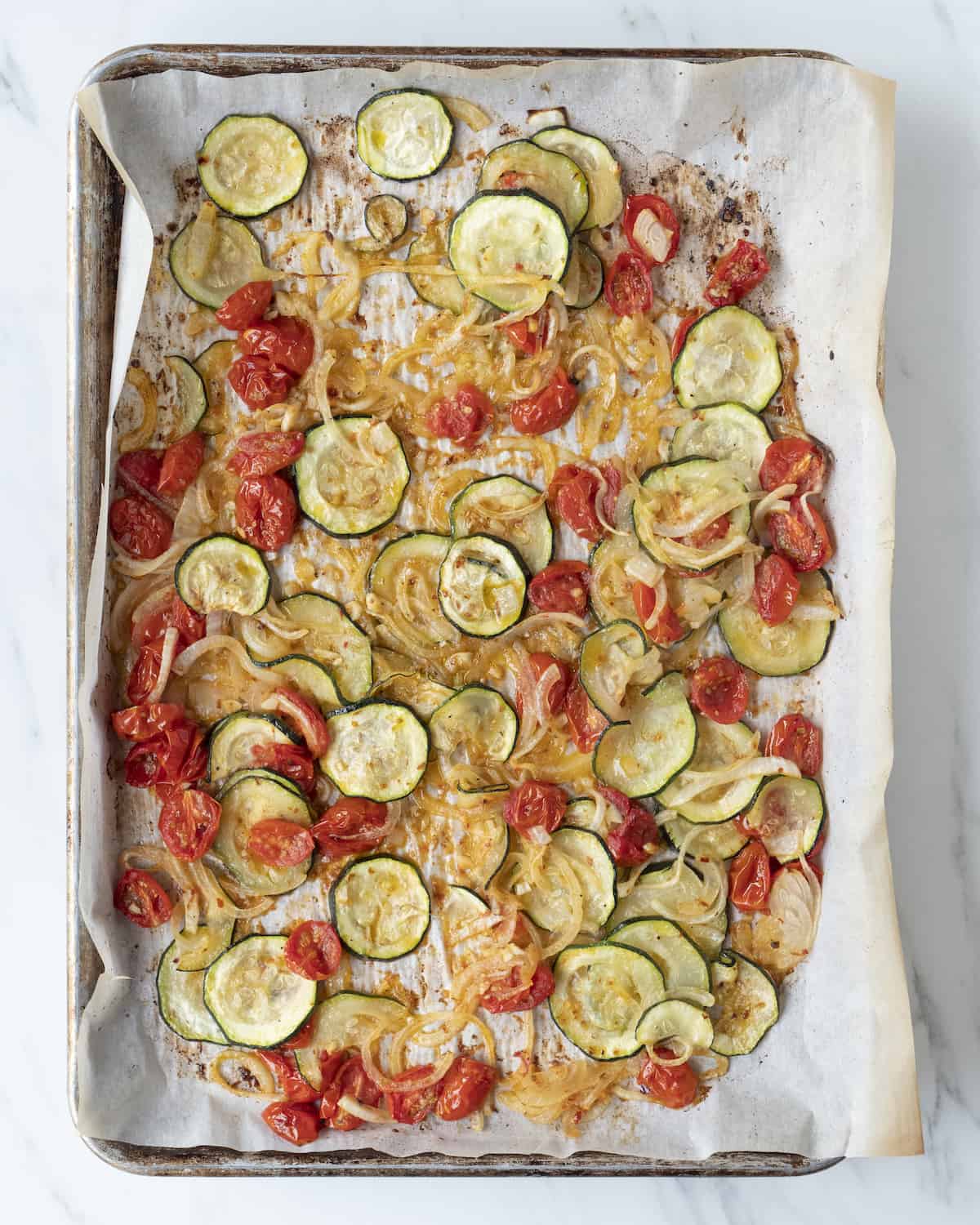 A sheet pan lined with parchment paper of cooked sliced zucchini, yellow onion, cherry tomatoes, and drizzled with olive oil. 