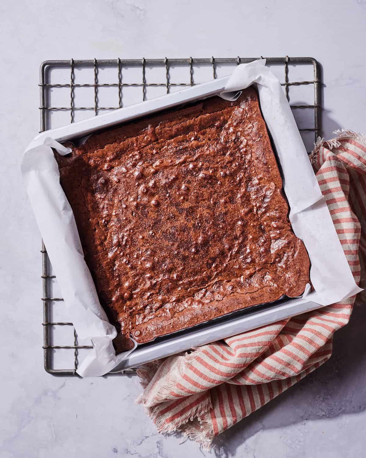 Freshly baked brownies in a 9x9 metal baking pan lined with parchment paper on a metal wire rack with an orange striped kitchen towel in the bottom right corner.