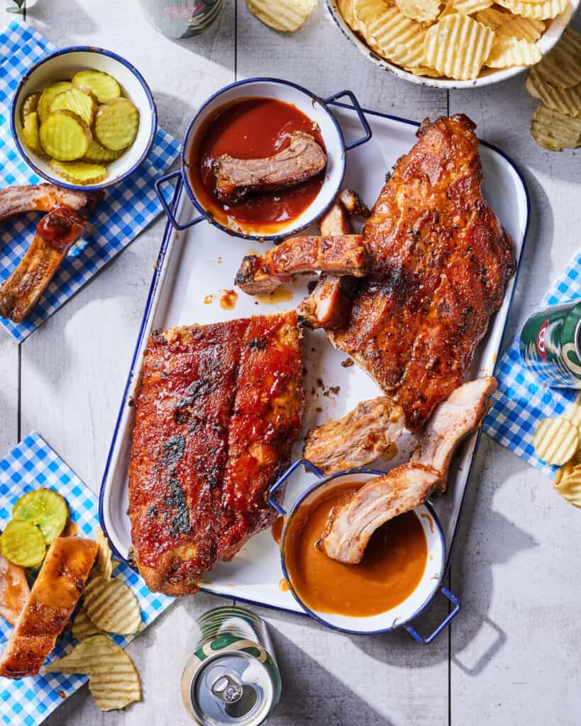 Rectangular platter with spiced bbq baby back ribs with a bowl of bbq sauce, pickles and cans of beer, surrounded with tissues being used to place the ribs, pickles and chips to eat.