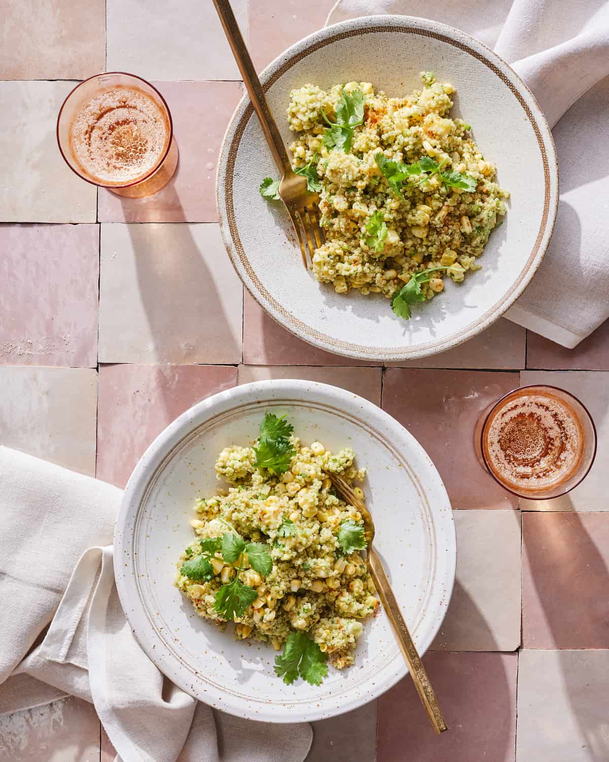 Two bowls of quinoa with roasted corn, avocado and and cilantro sitting next to two glasses of rose on a tile table.  
