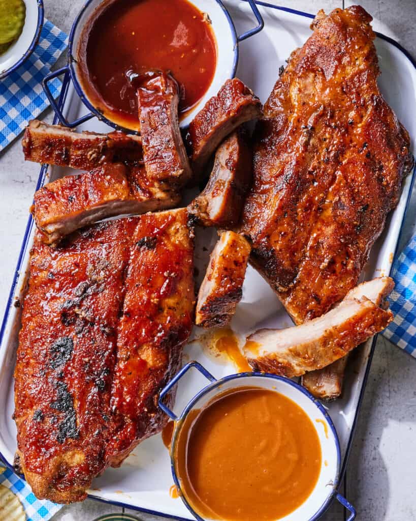 A rectangular platter with spiced BBQ baby back ribs cut up, with a bowl of bbq sauce.