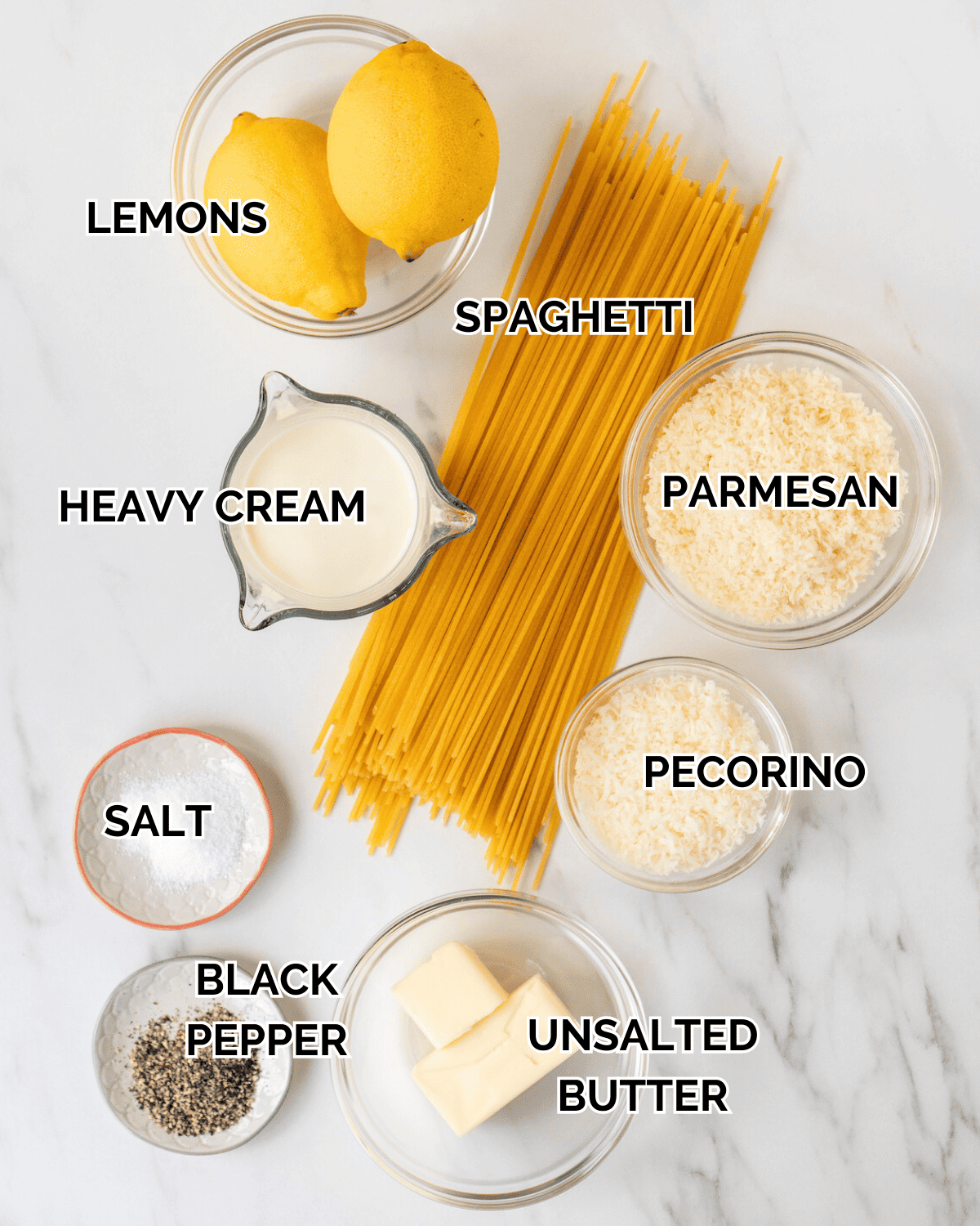 An overhead shot of all the ingredients in individual bowls.  Ingredients consist of 2 lemons, heavy cream, spaghetti, parmesan, salt, pecorino, black pepper, and unsalted butter.  