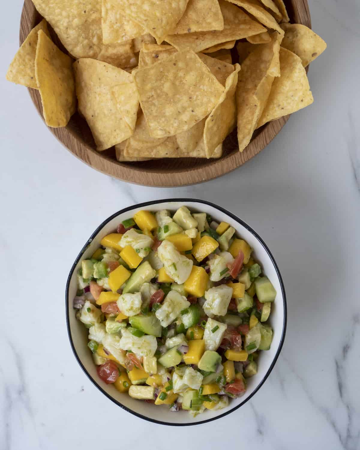 A wooden bowl filled with tortilla chips above a ceramic bowl with cubed mango, cucumber, avocado, red onion, tomato, jalapeño, chives, sea bass, and seasoned with salt, pepper, and lime juice. 