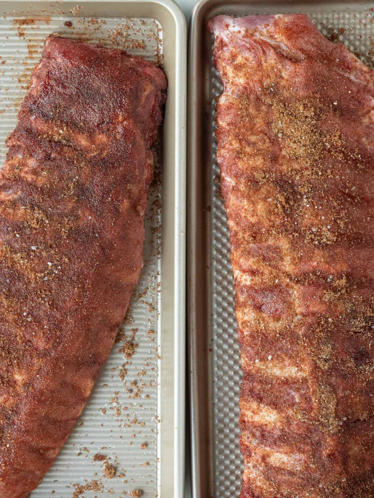 Two baking sheets, each with a rack of baby back ribs sprinkled with the spice mix all over.