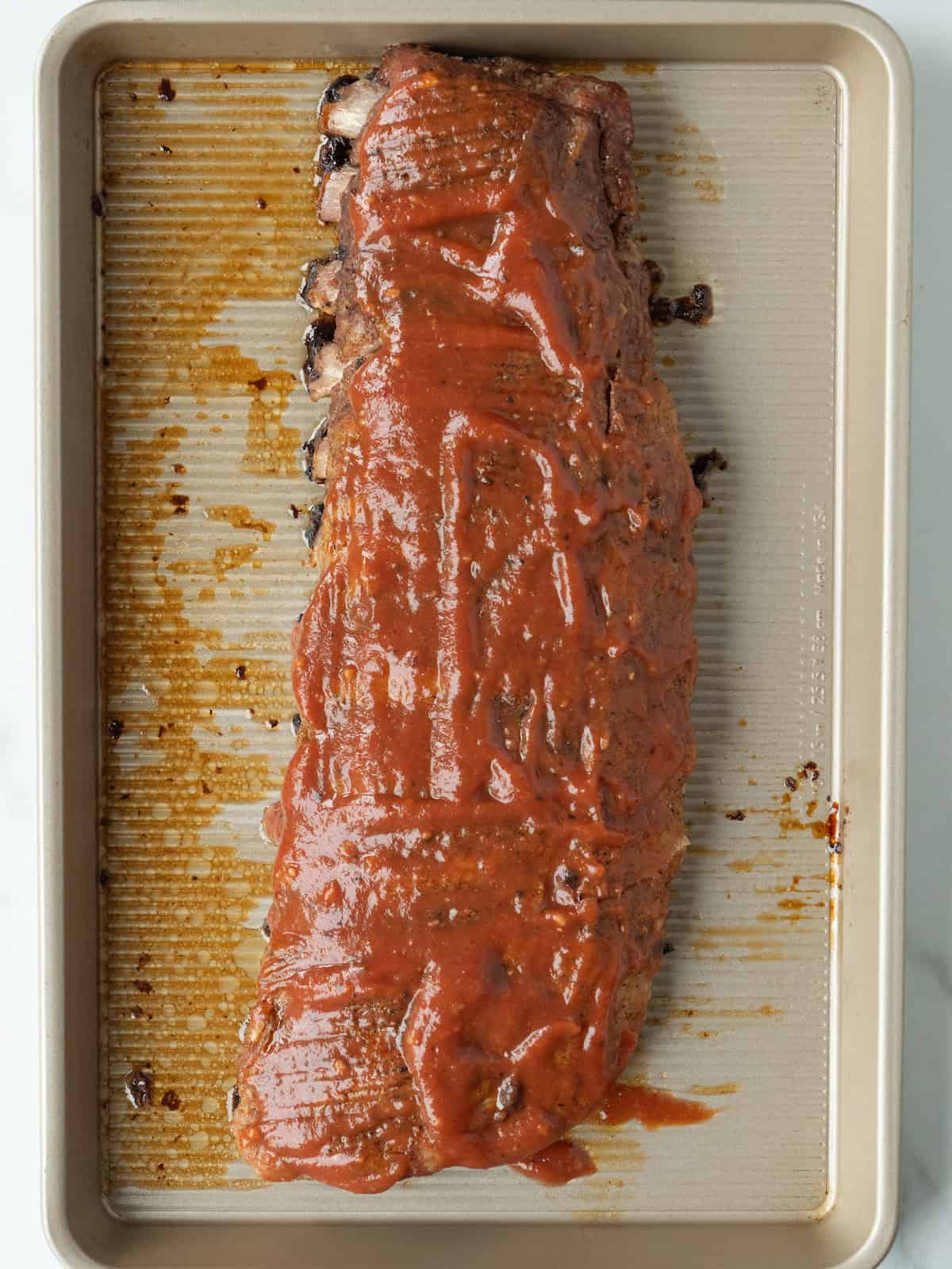 A baking sheet with a rack of baby back ribs brushed liberally with barbecue sauce.