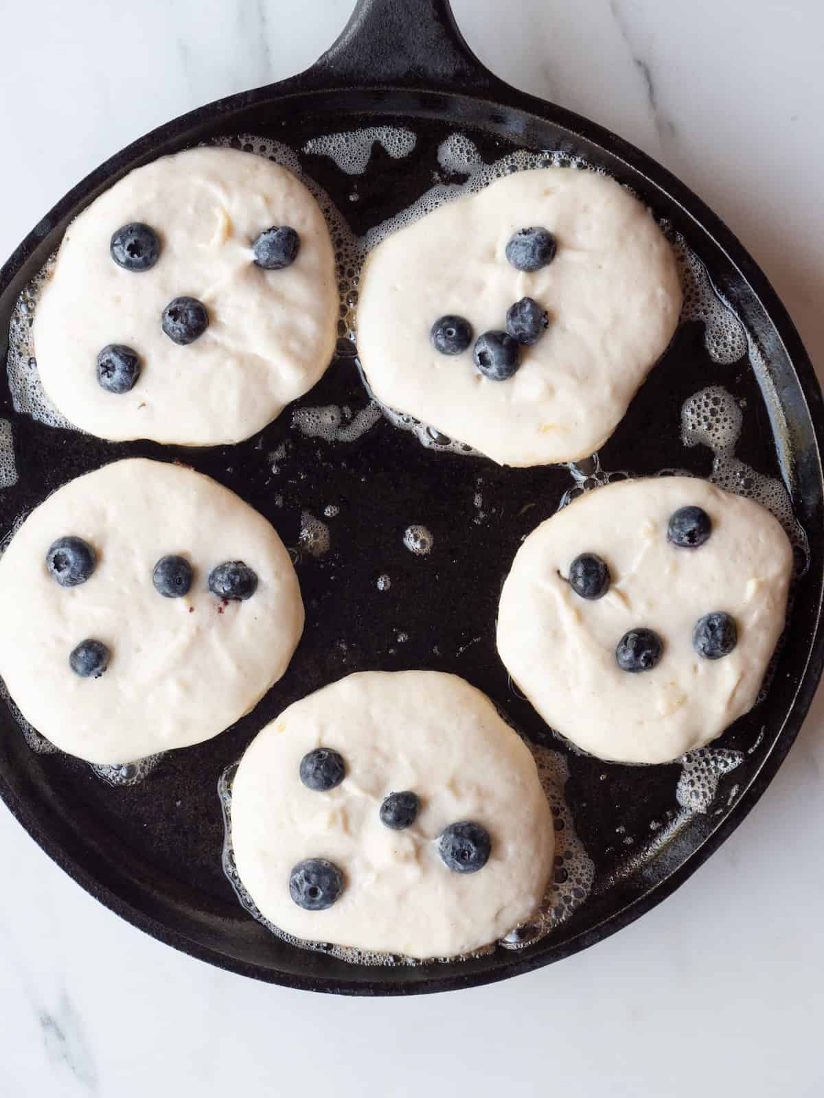 A pan with five small pancakes being cooked, topped with blueberries.