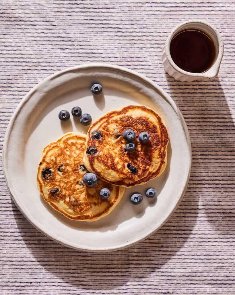 A plate with two blueberry buttermilk pancakes, topped with a few fresh blueberries, along with some maple syrup on the side.