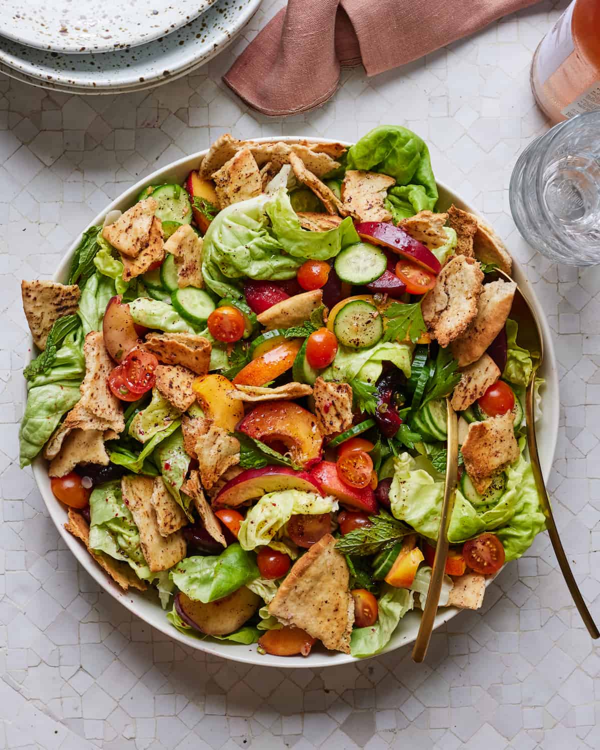 A large salad bowl filled with lettuce, halved cherry tomatoes, pita chips, sliced cucumbers, sliced stone fruit and mint.  