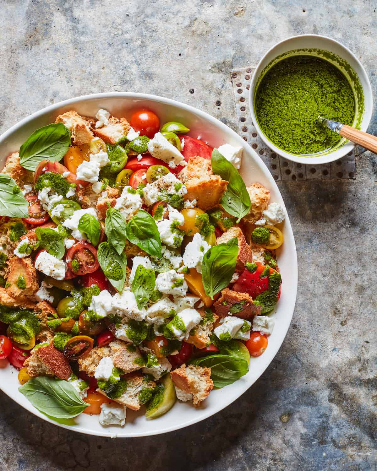 A giant salad bowl filled with croutons, chucks of mozzarella, halved tomatoes, and basil sitting next to a bowl of basil vin.